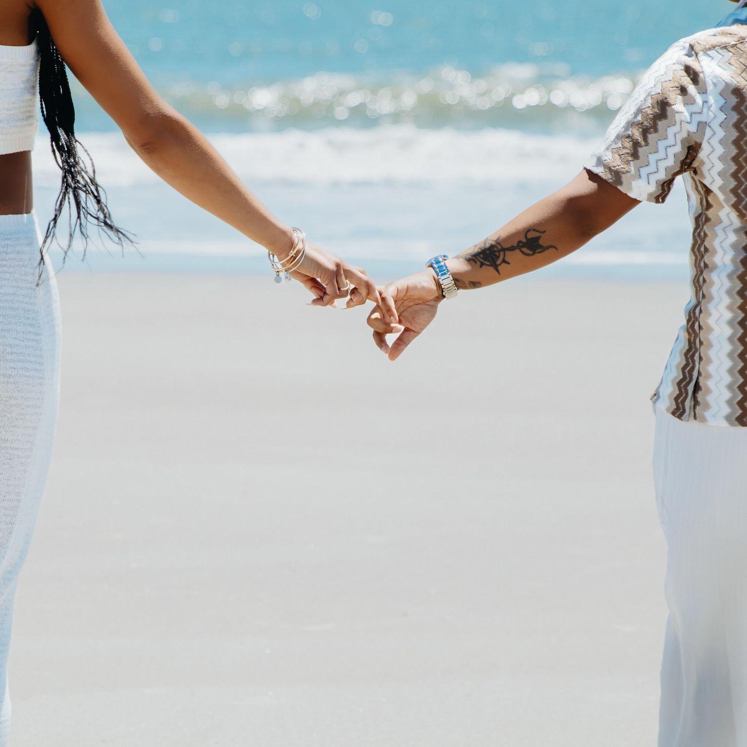 Couple holding hands on a sunny beach, showcasing tattoos and bracelets, dressed in white and patterned summer outfits.