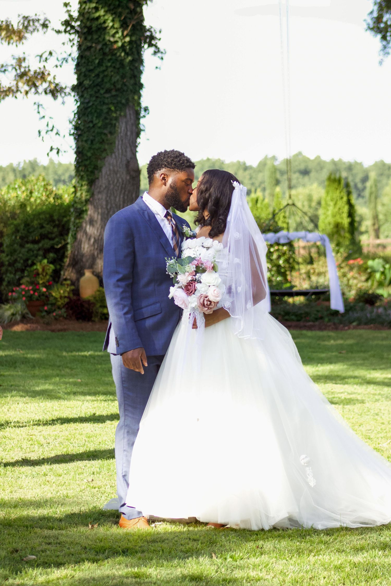 Wedding couple posing on lush green lawn with bride in flowing white dress and pink bouquet.