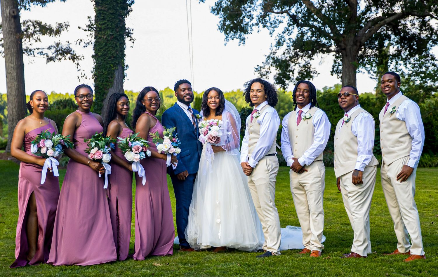 Wedding party in mauve and white attire poses for group photo outdoors under green trees.
