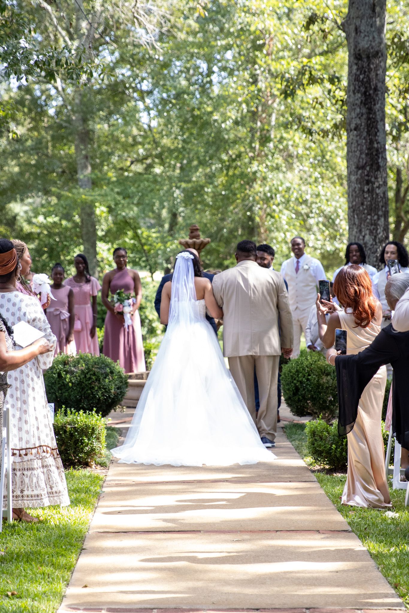 Wedding party walking along tree-lined pathway with bridesmaids in mauve dresses and white floral bouquets.