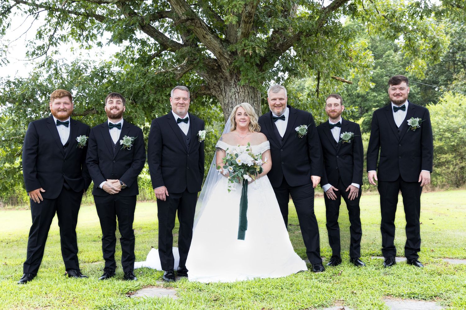 Bride in white gown poses with five groomsmen in black tuxedos outdoors under a large tree.