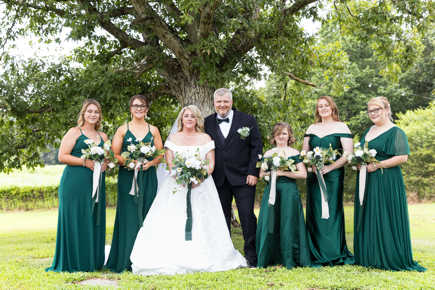 Wedding party poses outdoors under large tree, bride in white gown with bridesmaids in emerald green dresses holding white bouquets.