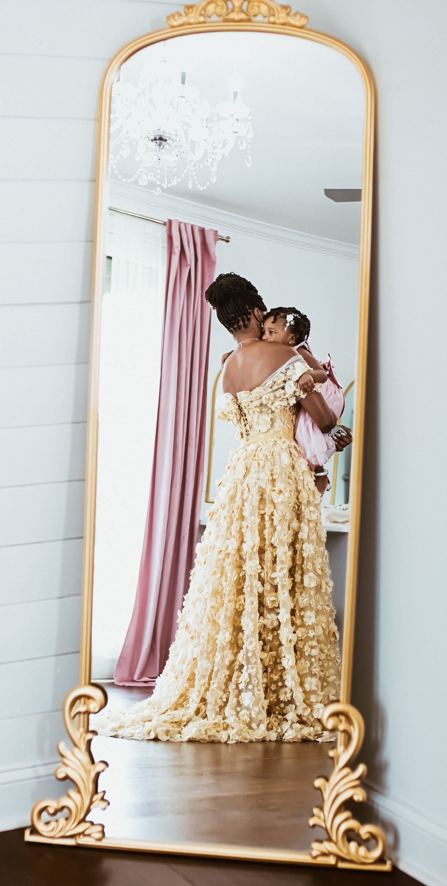 Ornate gold-framed mirror reflects an elegant cream-colored floral dress with flowing fabric in a light-filled room.