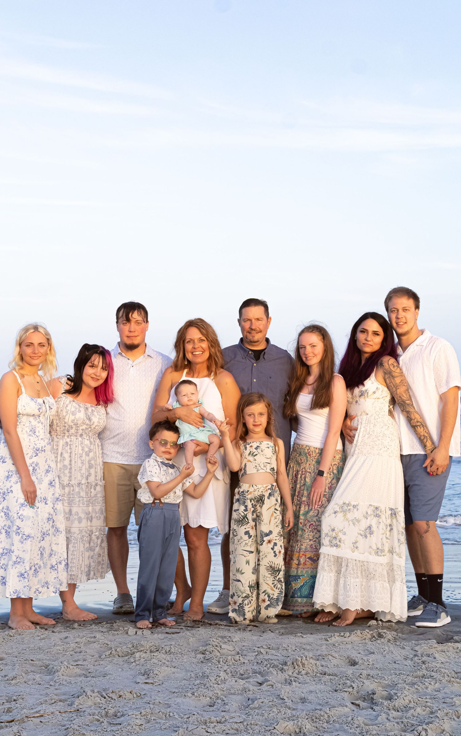 Large family group photo on a sandy beach at sunset, featuring adults and children posing together by the ocean.