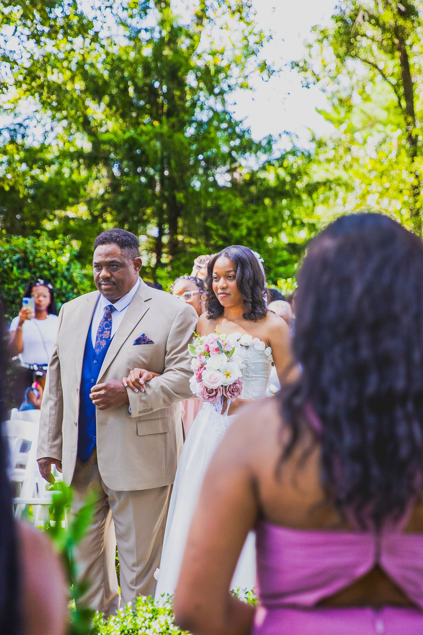 Bride in white gown walks down outdoor aisle with escort in tan suit at a sunny garden wedding ceremony.
