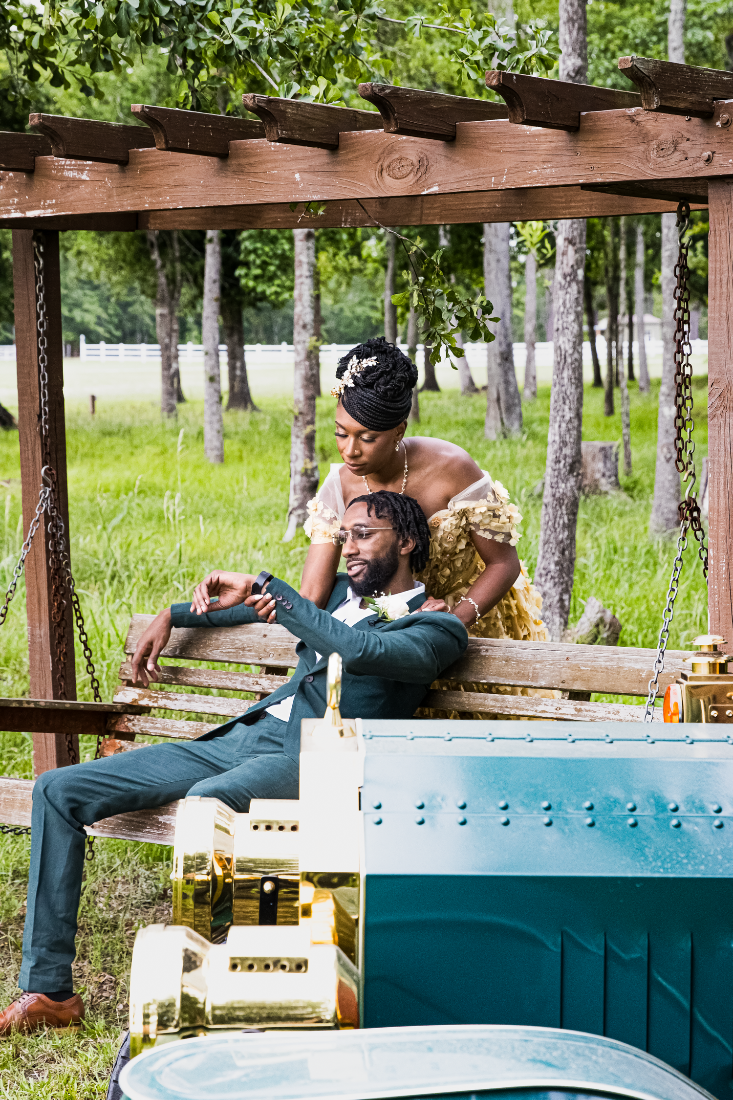 A couple in elegant attire poses under a wooden pergola on a swing bench, surrounded by lush greenery.