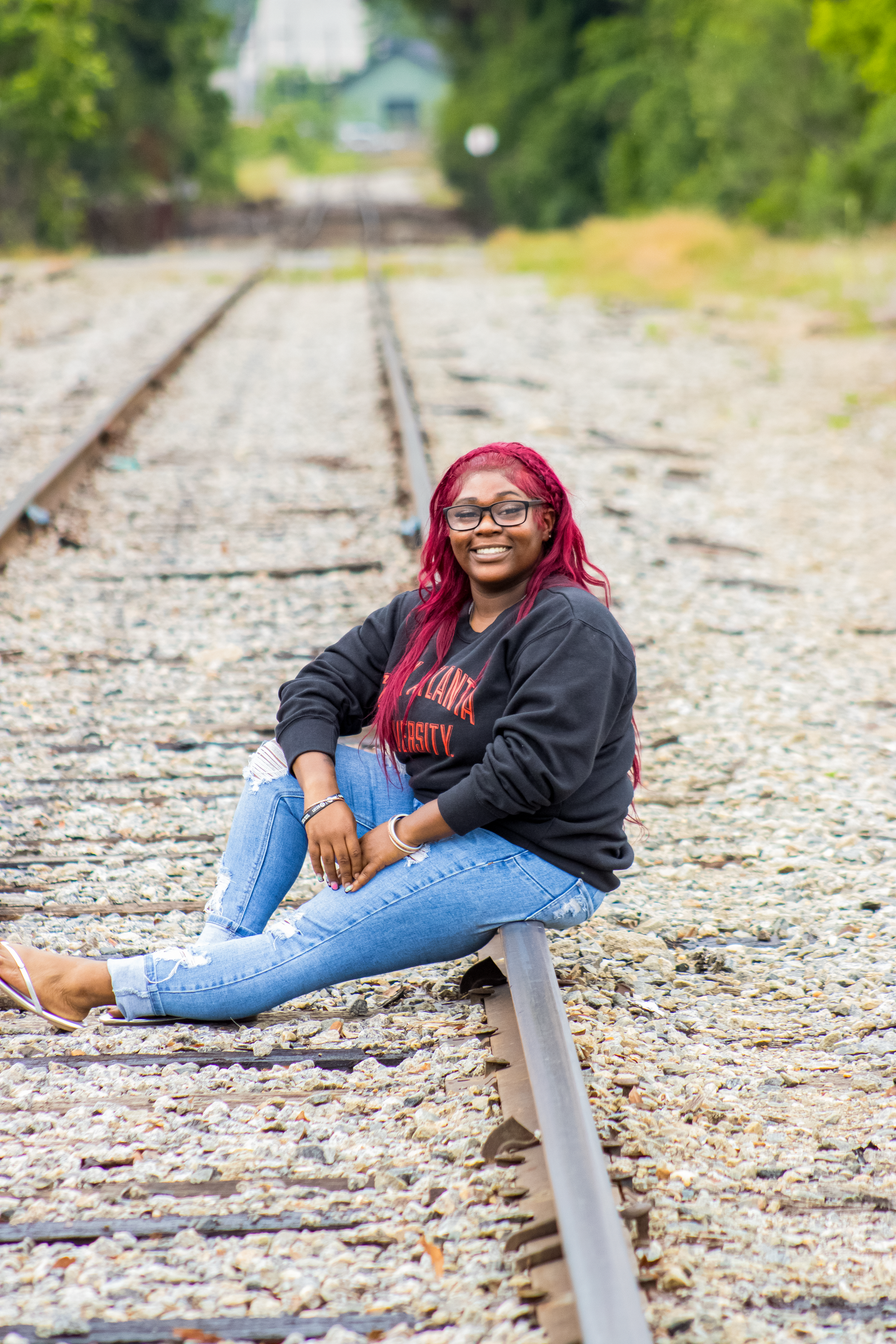 Person posing casually on railroad tracks wearing red hair, glasses, and black hoodie.