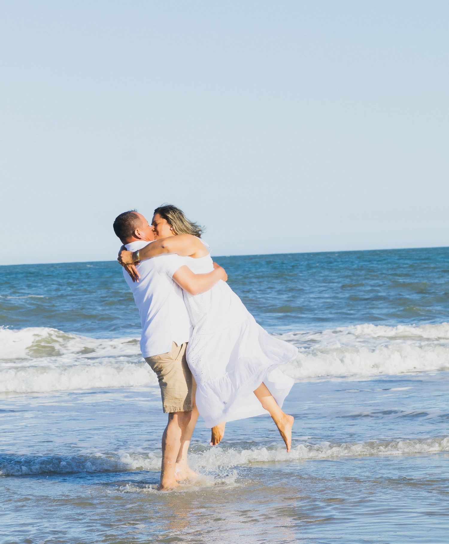 Couple sharing a romantic kiss on the beach, man lifting woman in white dress with ocean waves behind them.