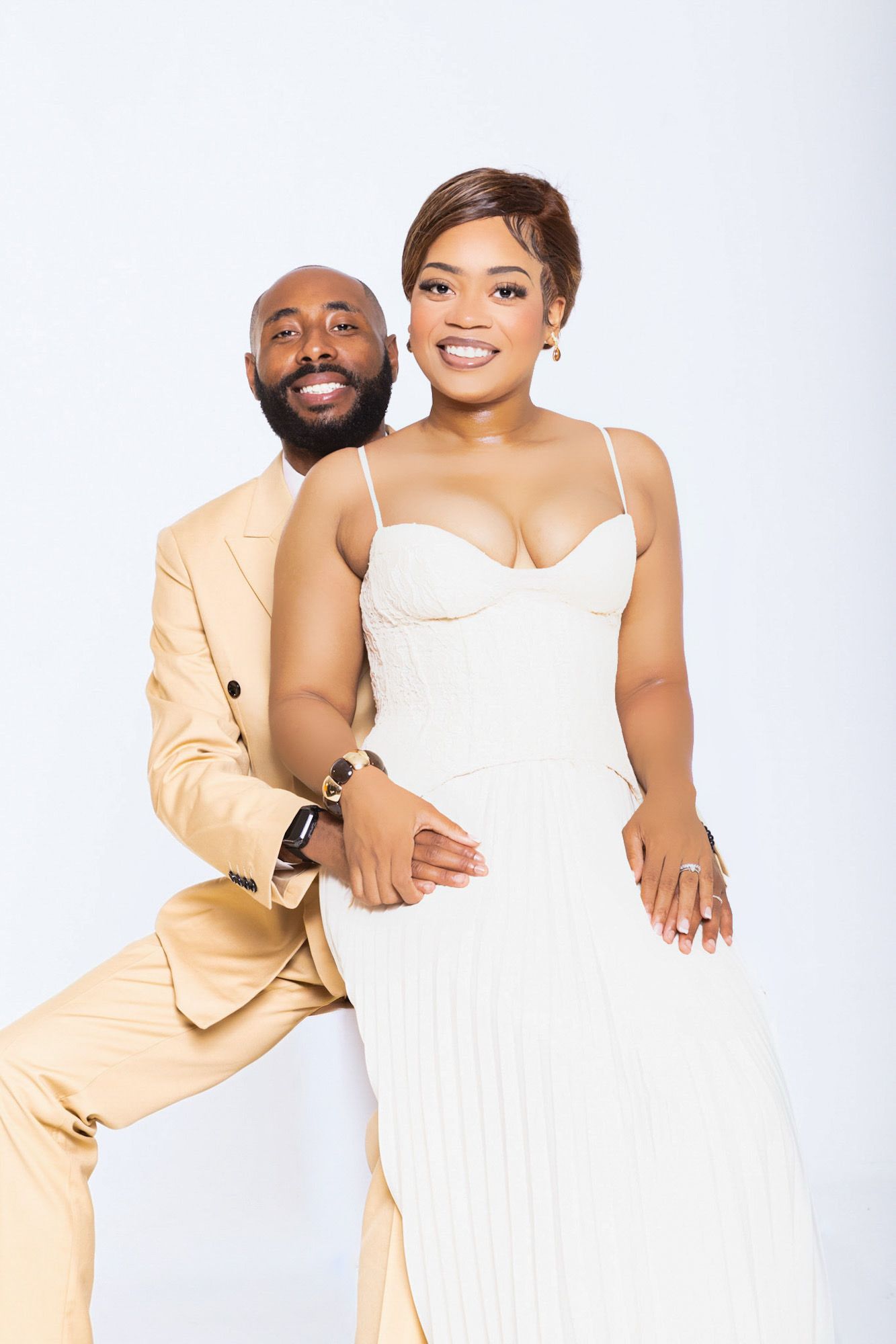 Elegant couple posing in studio, man in beige suit embracing woman in white spaghetti-strap dress against white background.