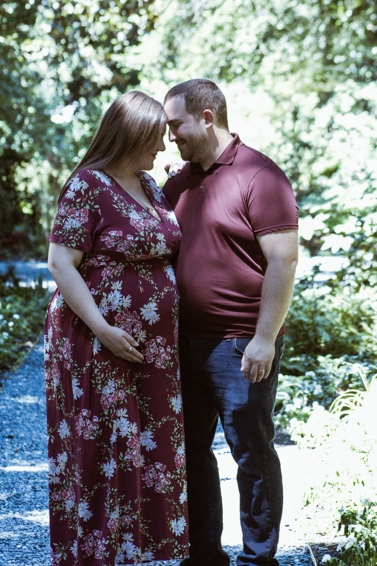 Expecting couple shares intimate moment on forest path wearing burgundy floral dress.