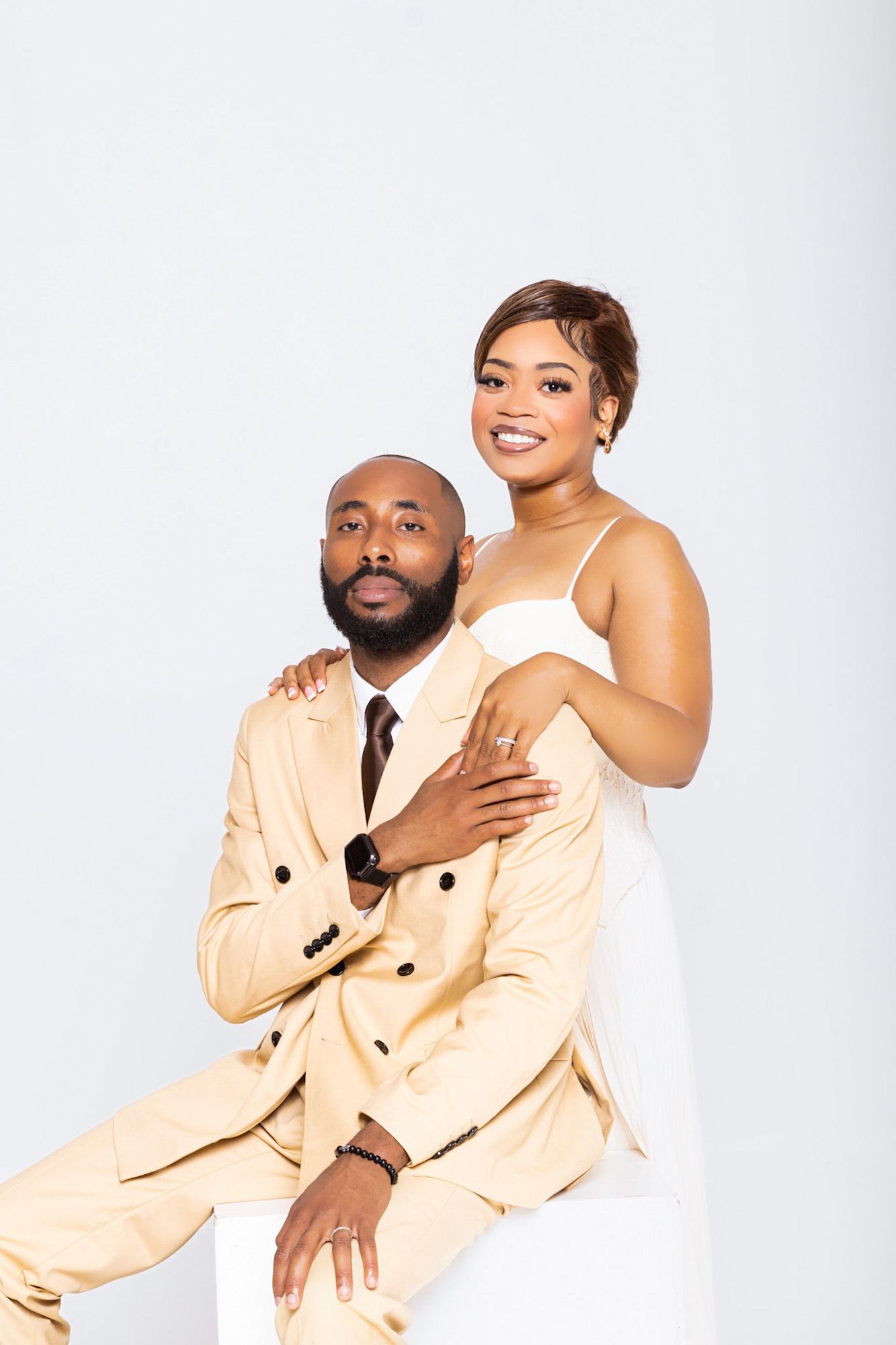 African American couple in wedding attire, groom in beige suit seated, bride in white dress smiling, studio portrait.