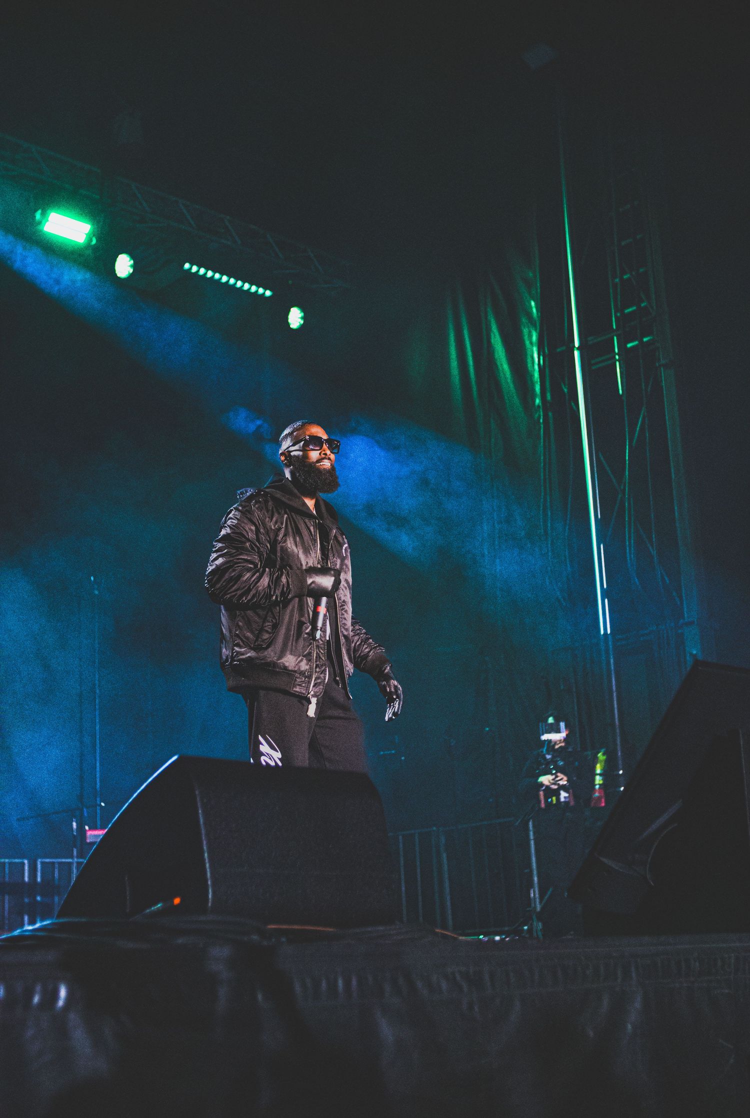 A performer in dark clothing stands on stage against a dramatic blue and green illuminated backdrop during a concert.