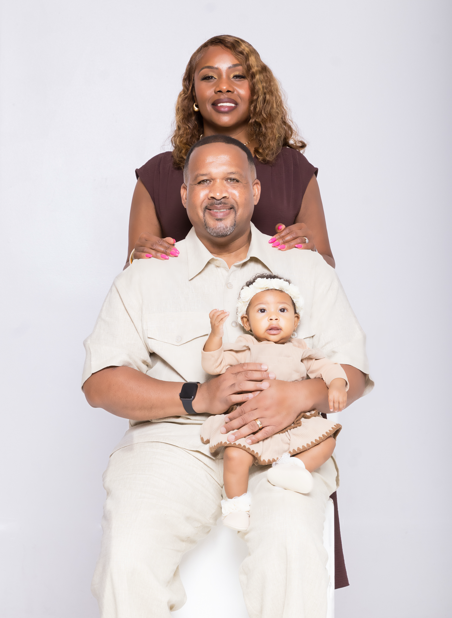 A family portrait featuring a man holding a baby, with a woman standing behind them, smiling warmly.