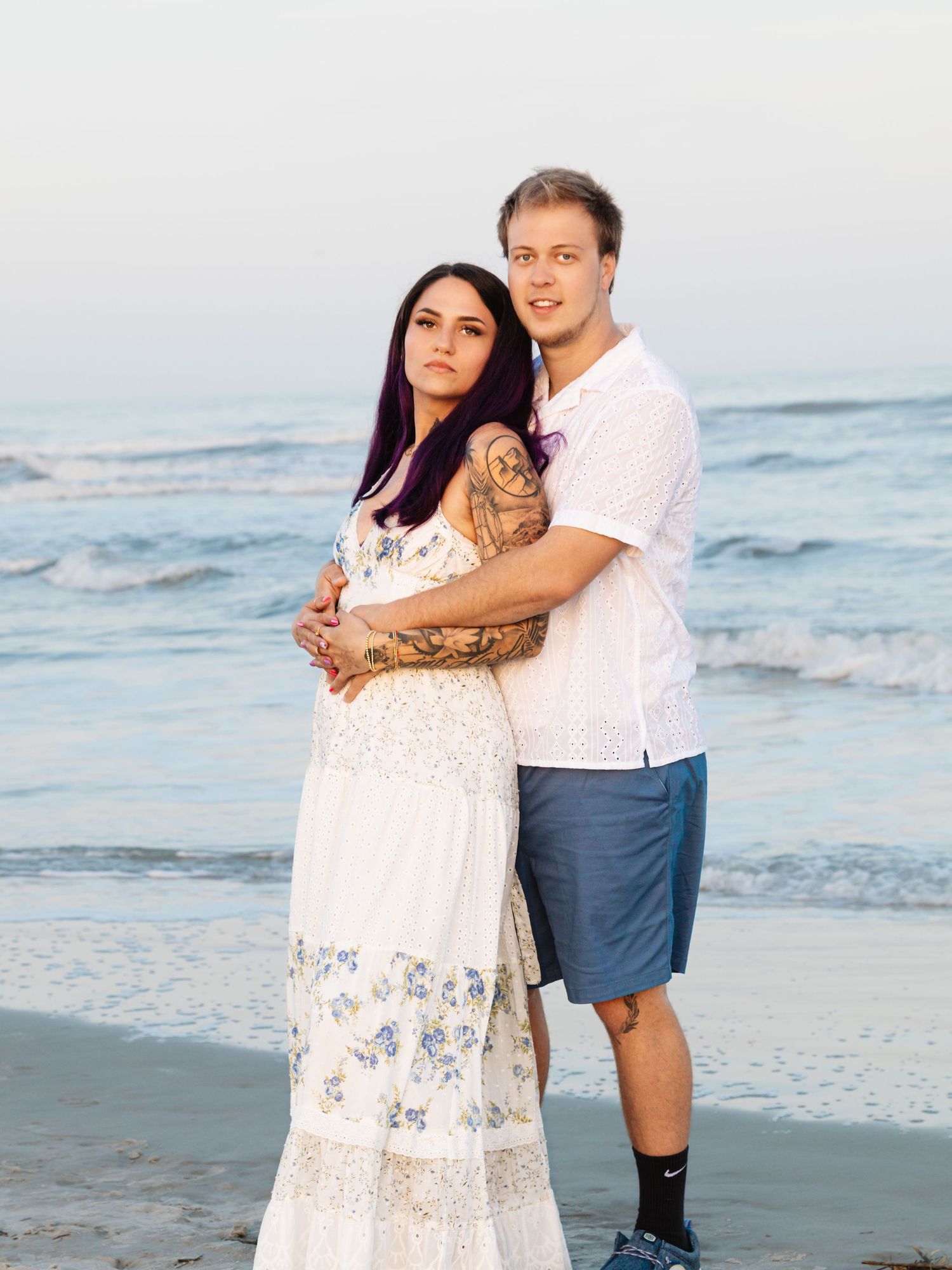 A couple embraces on a beach, woman in white floral dress with purple hair and tattoos, man in white shirt and blue shorts.