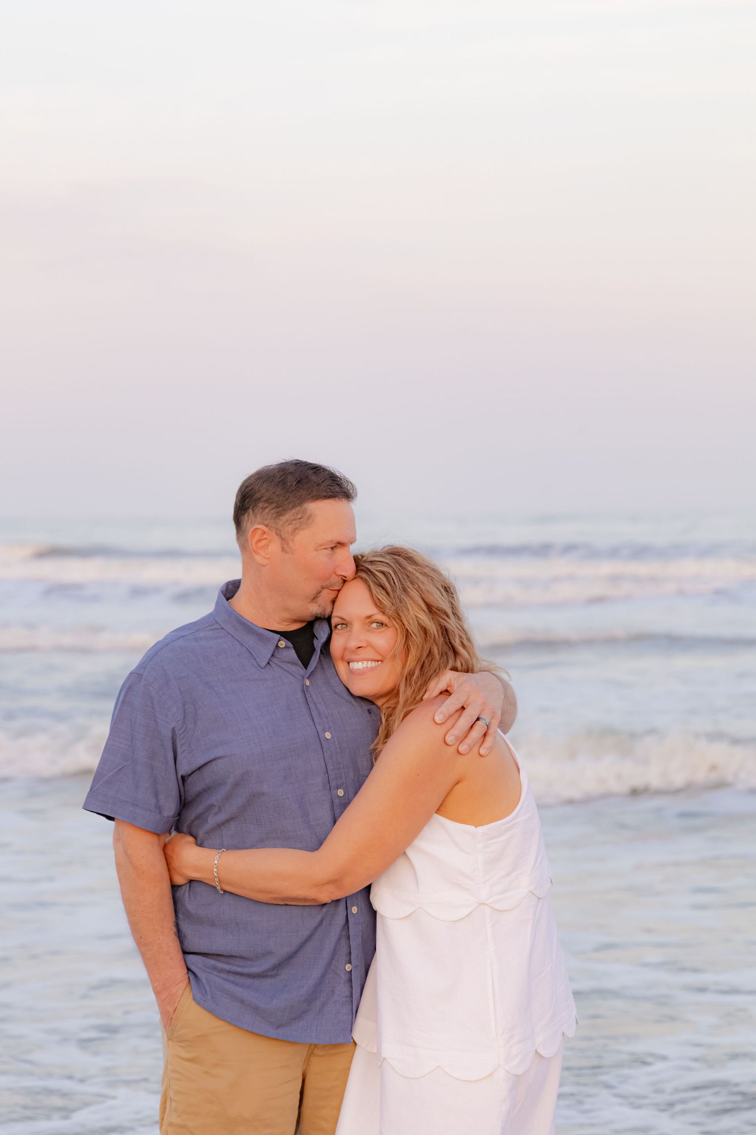 Couple embracing on beach at sunset, man kissing woman's head, waves in background.