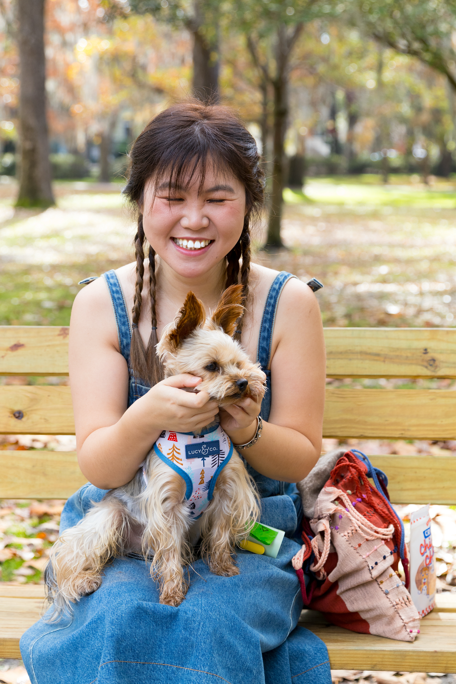 A joyful person in denim overalls sits on a park bench with a small Yorkshire Terrier dog on a sunny autumn day.