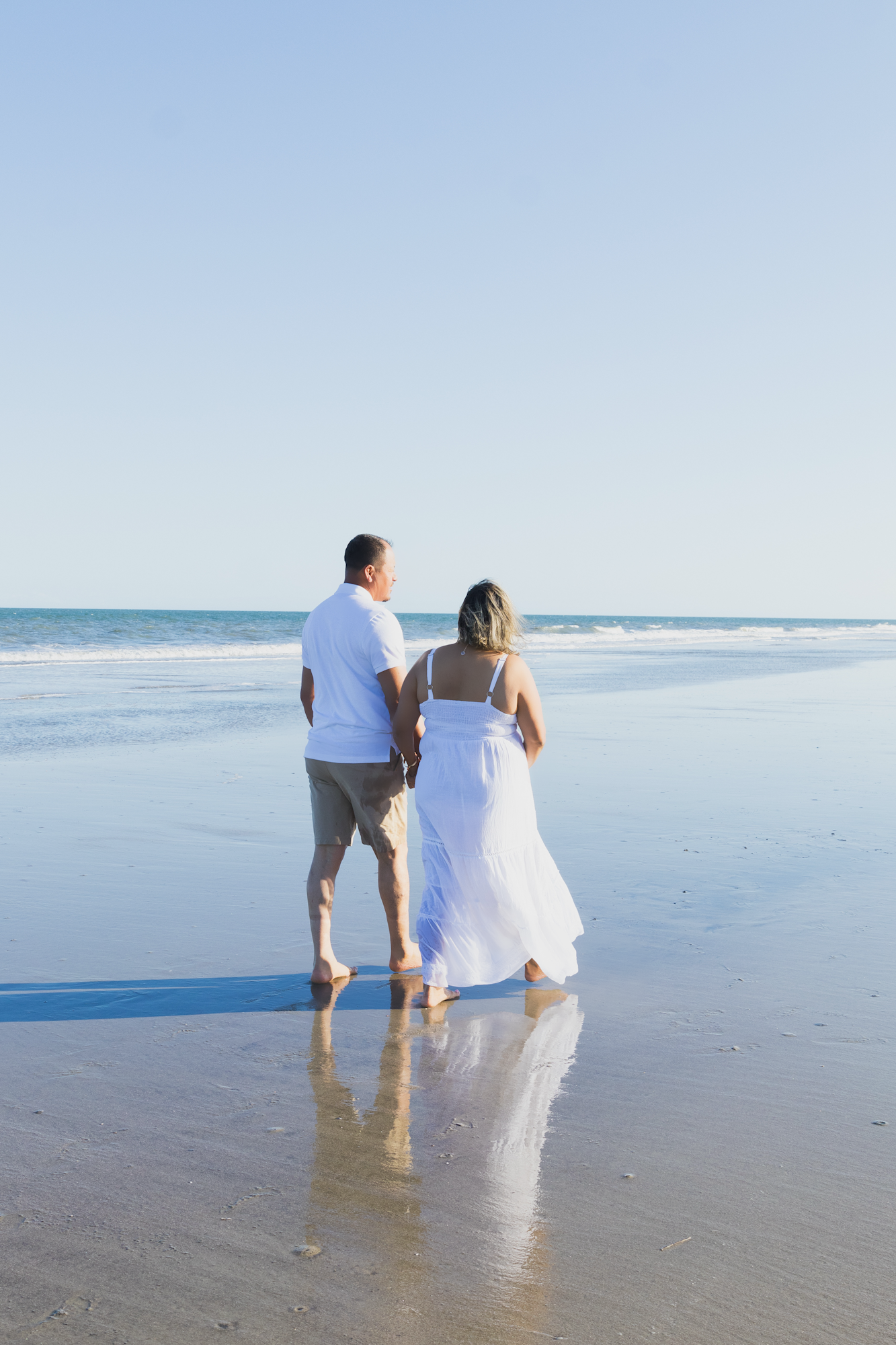 Couple in white holding hands walking barefoot on a reflective wet beach toward the ocean on a clear sunny day.