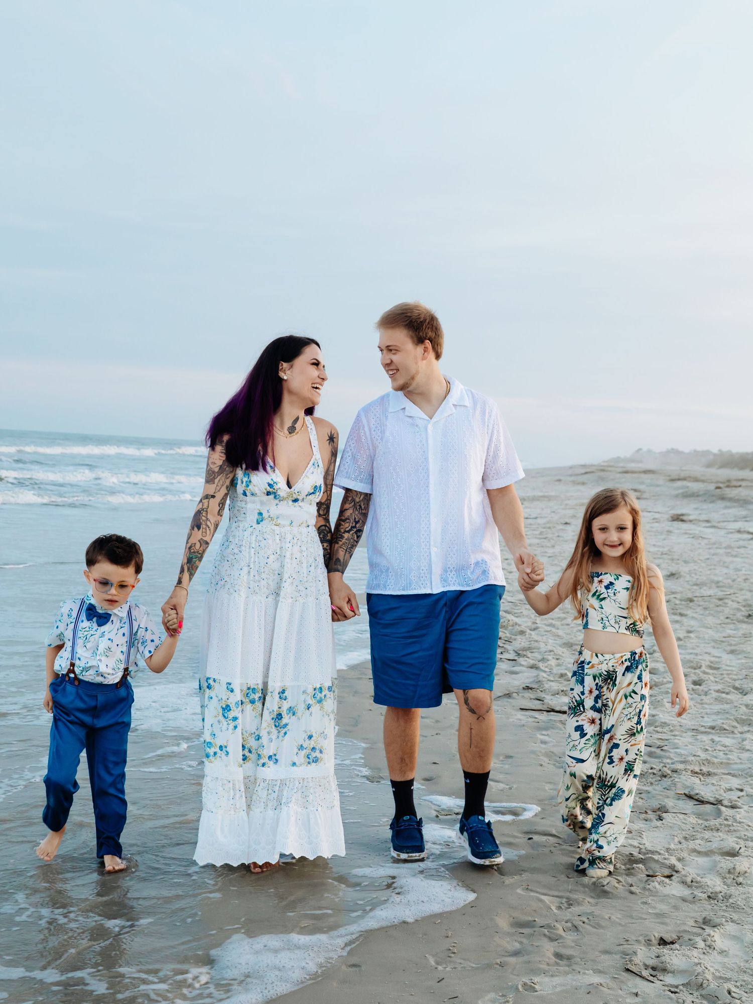 Family of four walking on beach, parents holding children's hands, waves in background, wearing coordinated blue and white outfits.