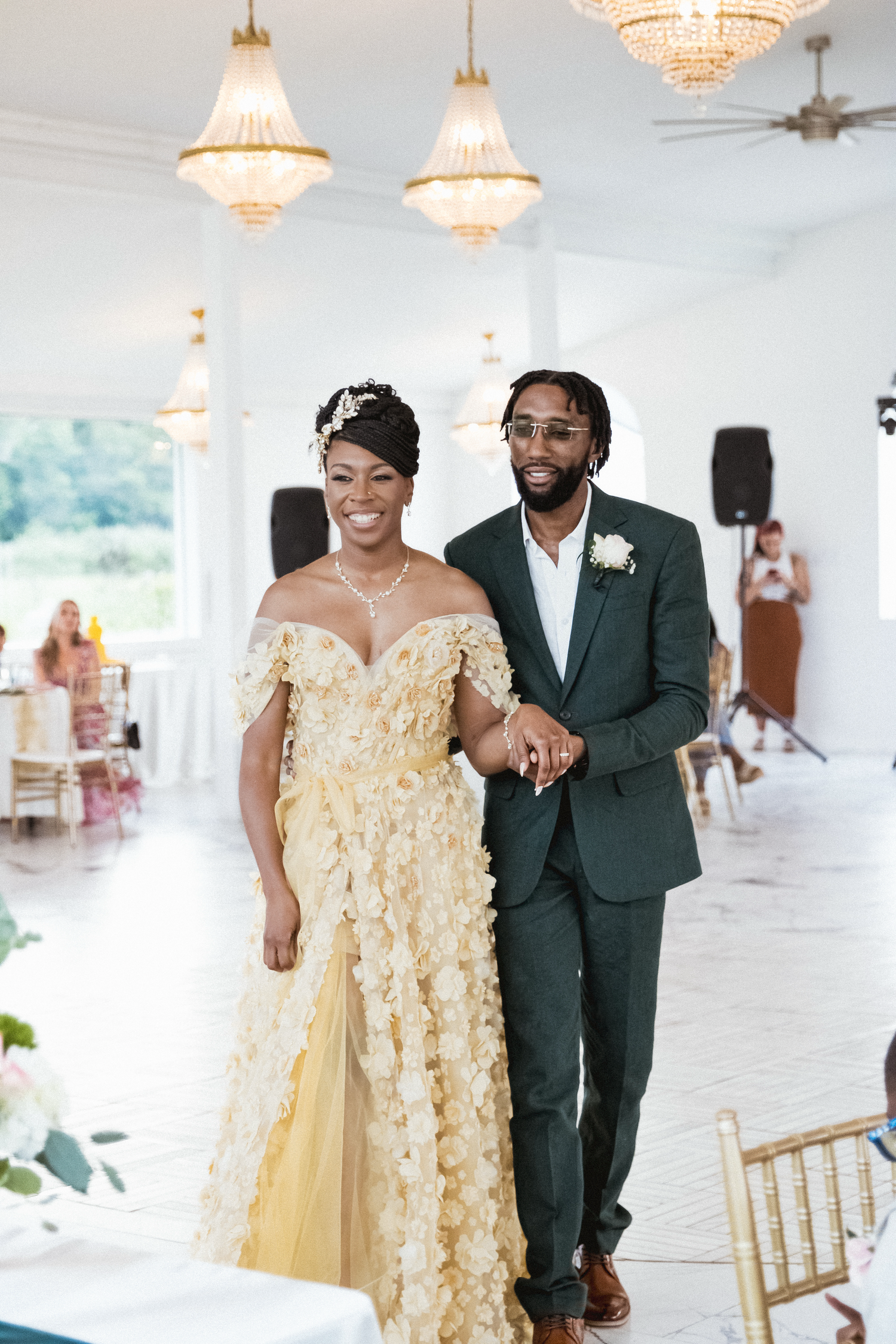 A couple in a yellow gown and dark suit walk together in an elegant event space with crystal chandeliers.