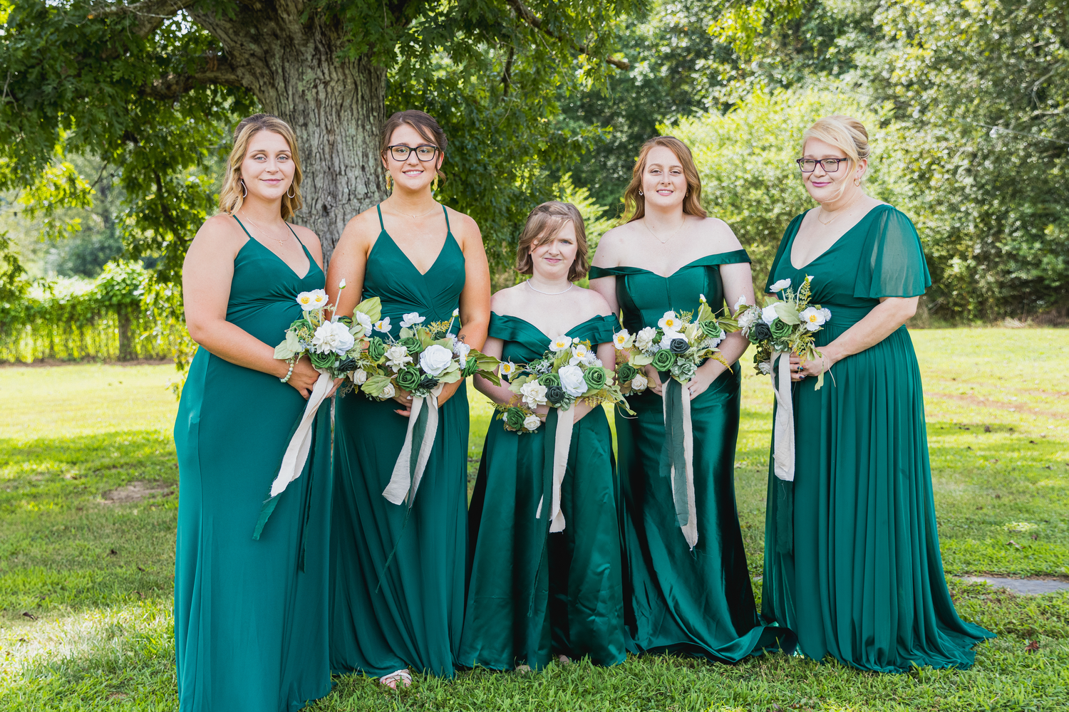 Five bridesmaids in emerald green gowns holding floral bouquets pose outdoors on a green lawn near a large tree.