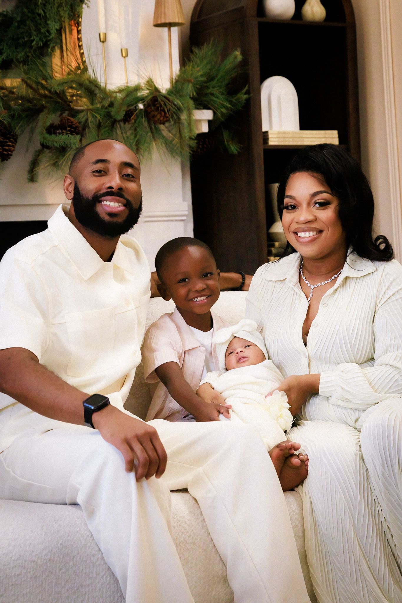 A family wearing white sits together on a couch with holiday greenery decorations visible in the background.