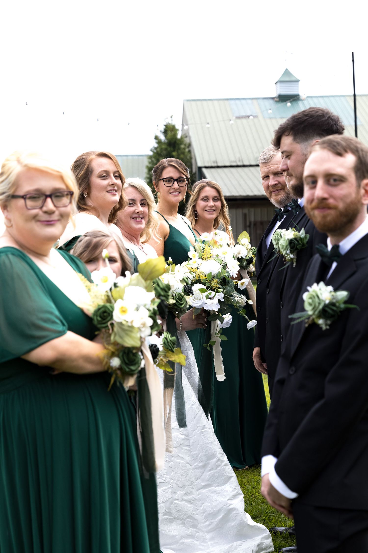 Wedding party in green dresses and black tuxedos posing outdoors near a barn venue.