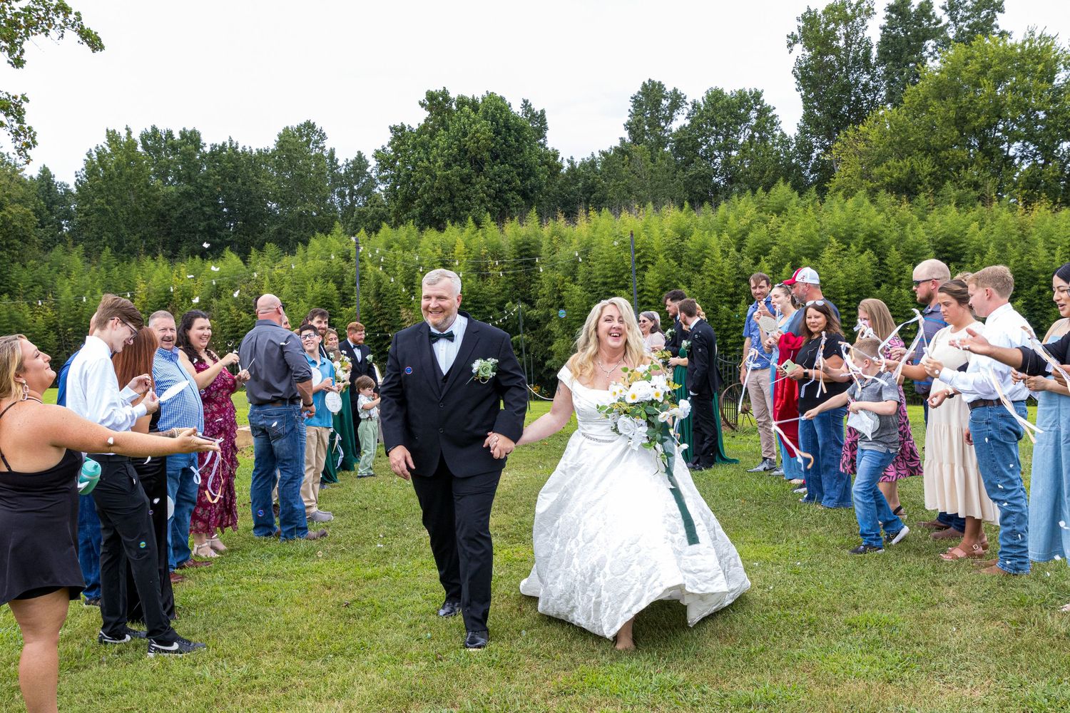 Bride and groom walk through cheering guests outdoors at a garden wedding ceremony surrounded by lush green trees.
