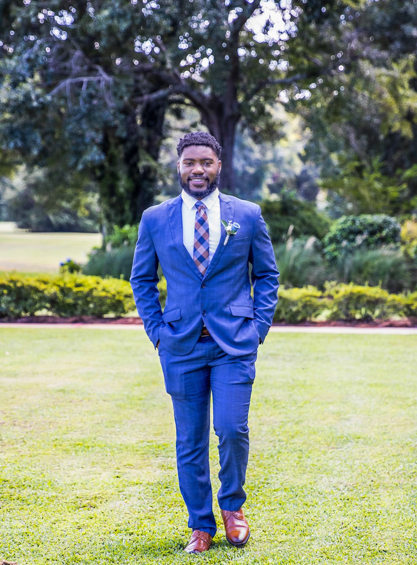 Man in blue suit with plaid tie and boutonniere standing on green lawn with trees and shrubs in background.
