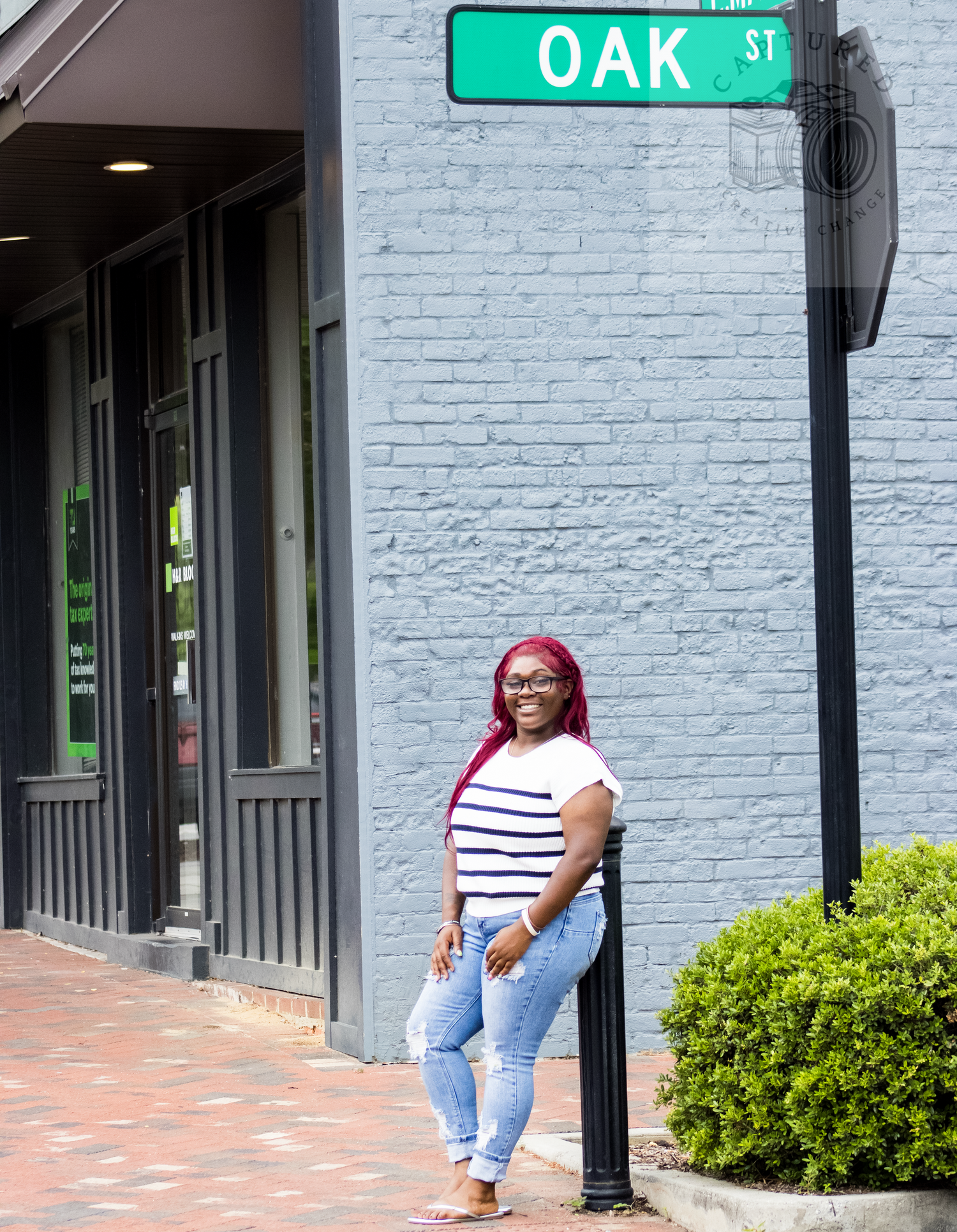 Woman with red hair poses by Oak St sign on brick sidewalk in casual striped top and jeans.