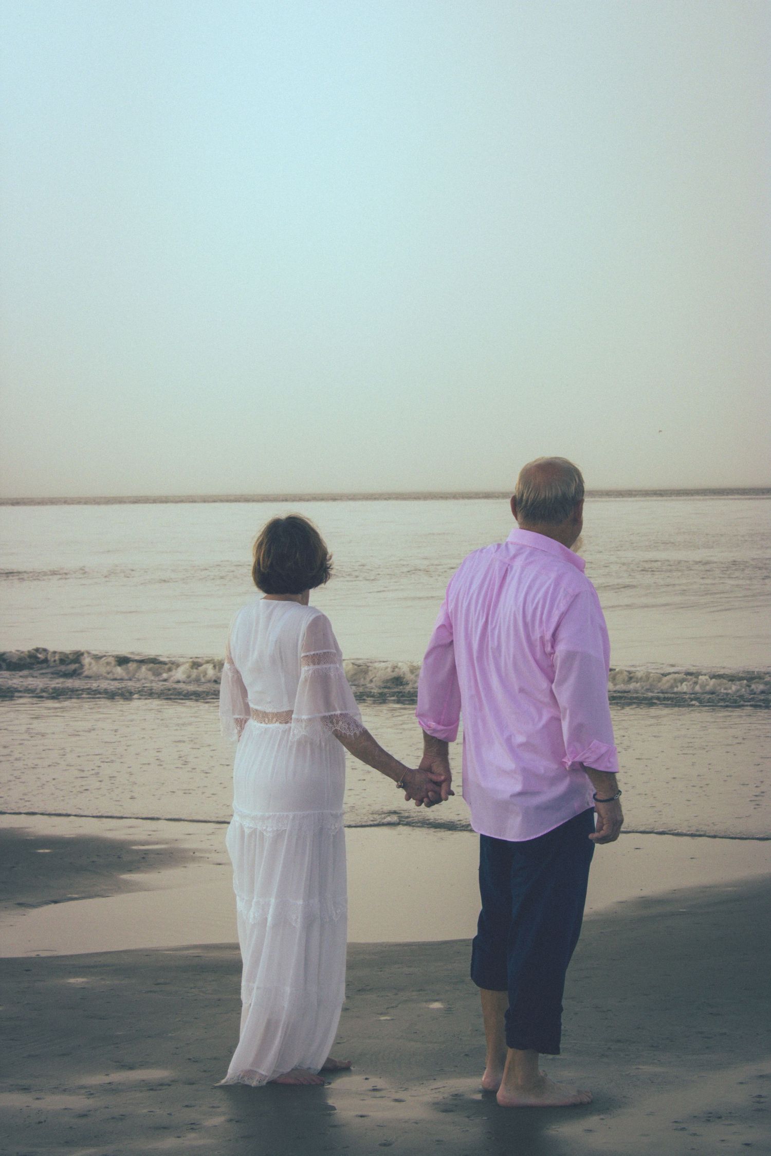 Couple holding hands while standing on beach at sunset in casual attire.