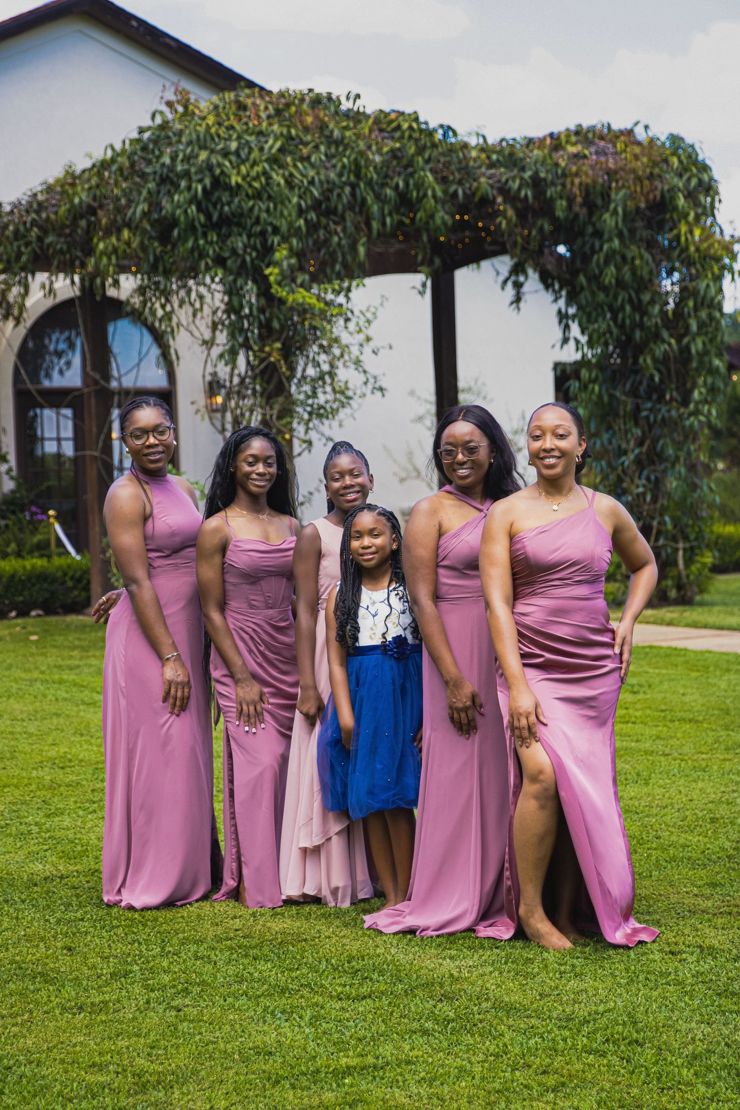 Bridal party wearing matching pink dresses poses on grass in front of a vine-covered building.