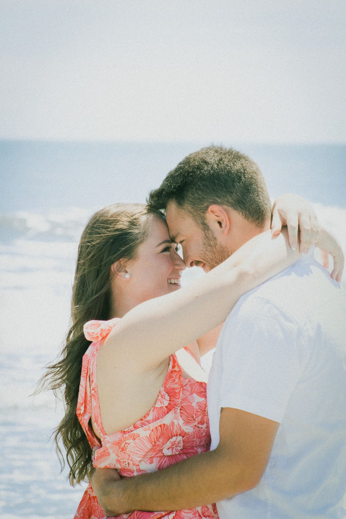 Couple embraces tenderly on a bright sunny beach with ocean in background.