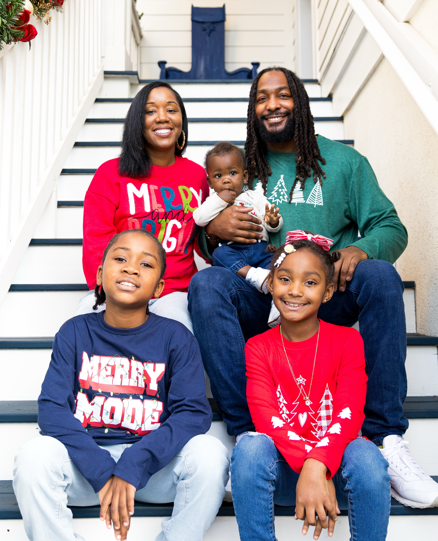 A family wearing red and blue holiday sweaters sits together on white stairs for a festive portrait.
