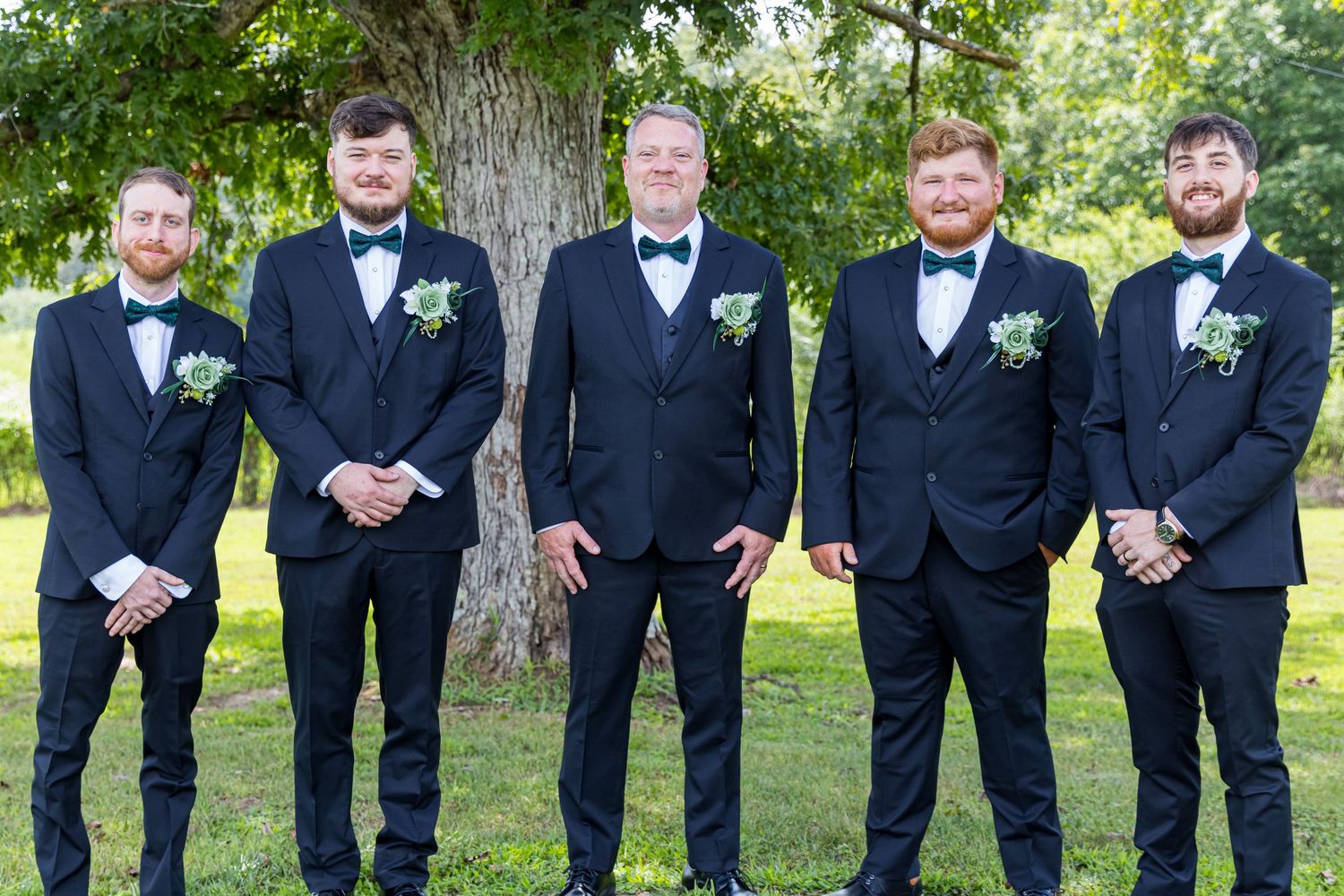 Five groomsmen in black tuxedos with boutonnieres pose together outdoors under a large tree.