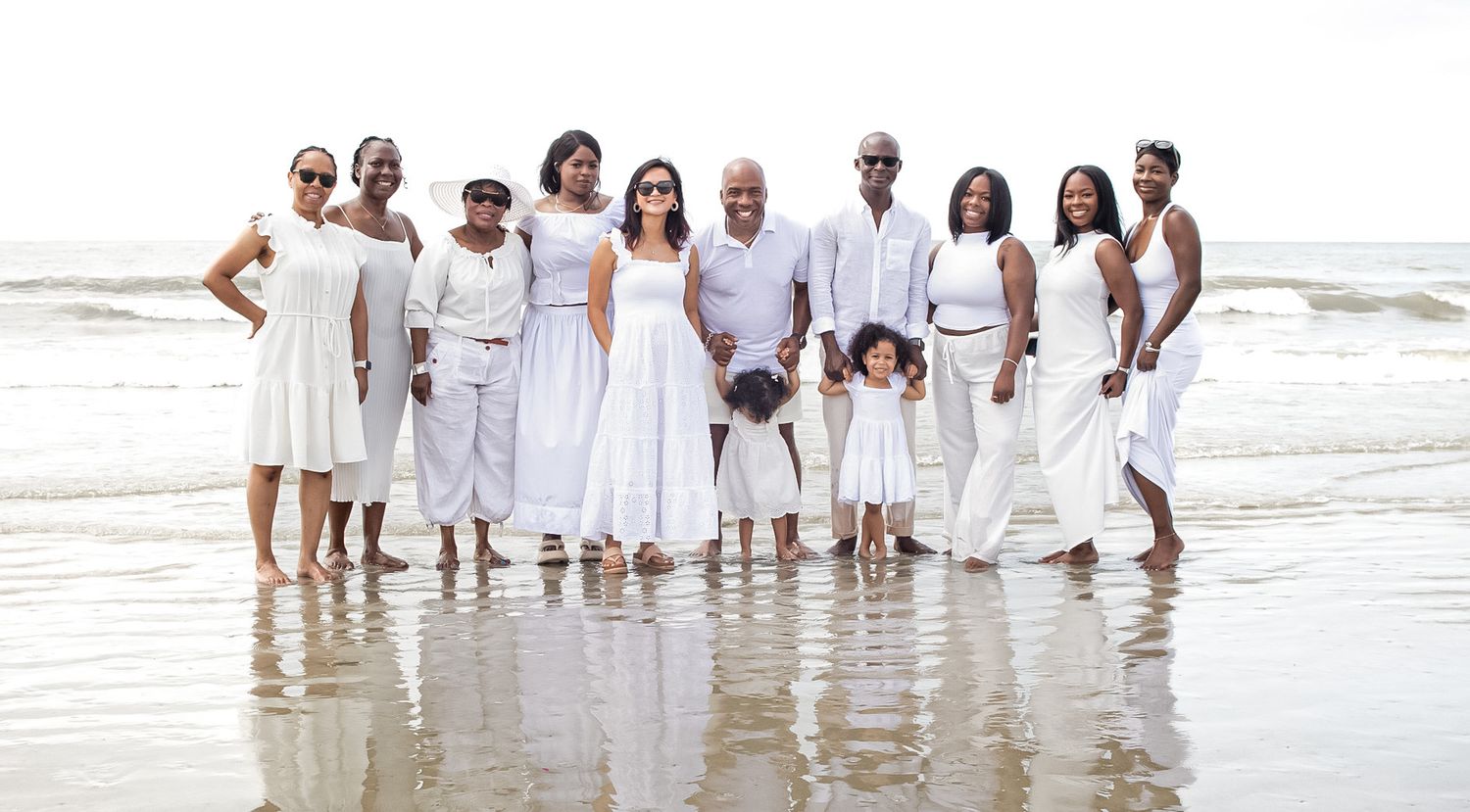A group in white outfits poses together for beach family photos along the shoreline at sunset.