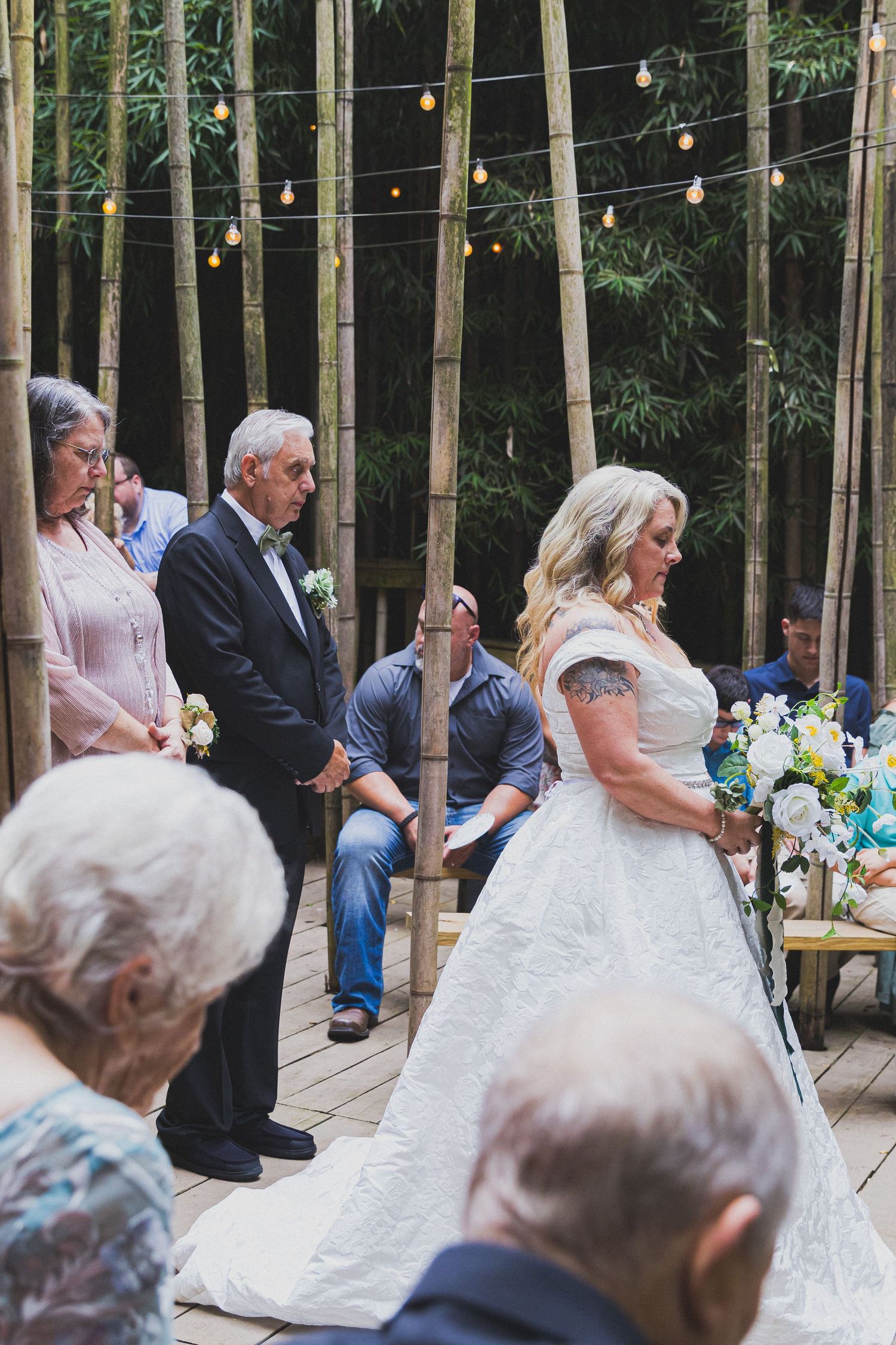 Bride in white lace gown stands at outdoor wedding ceremony surrounded by bamboo trees and string lights.