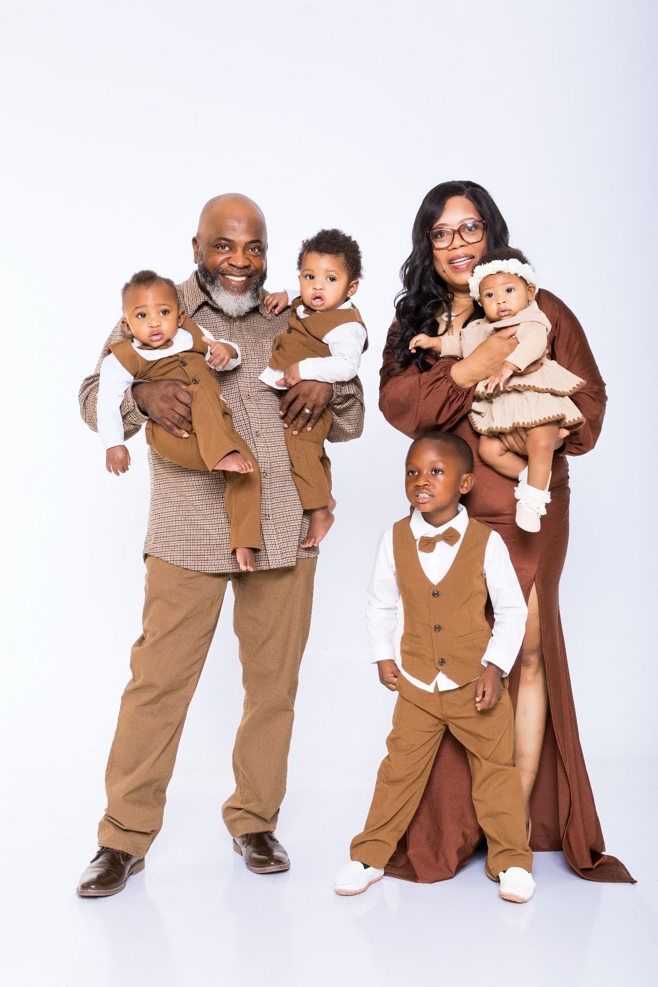 A joyful multigenerational Black family poses in coordinating brown outfits, with grandparents holding three young children.