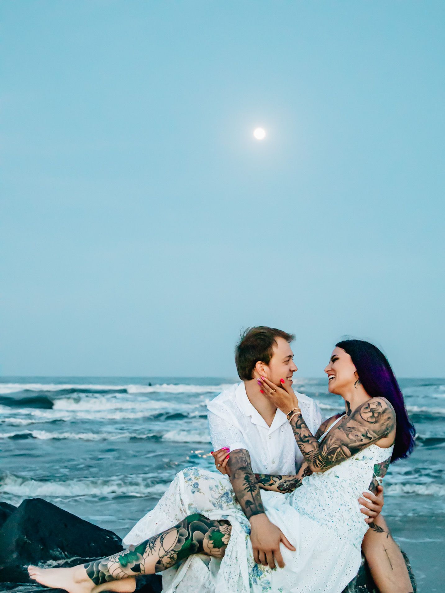 Tattooed couple embracing romantically on a beach at dusk with ocean waves and a full moon glowing in the blue sky.