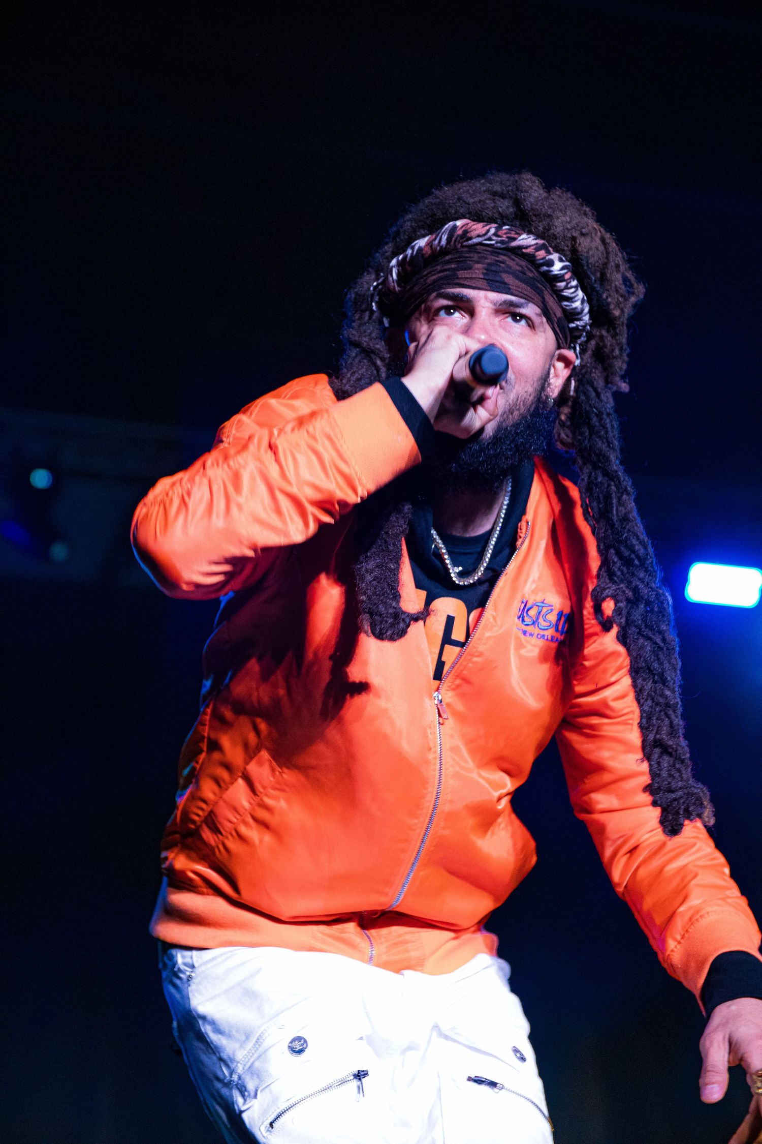 Performer in orange and white jacket singing passionately on stage with dramatic blue lighting.