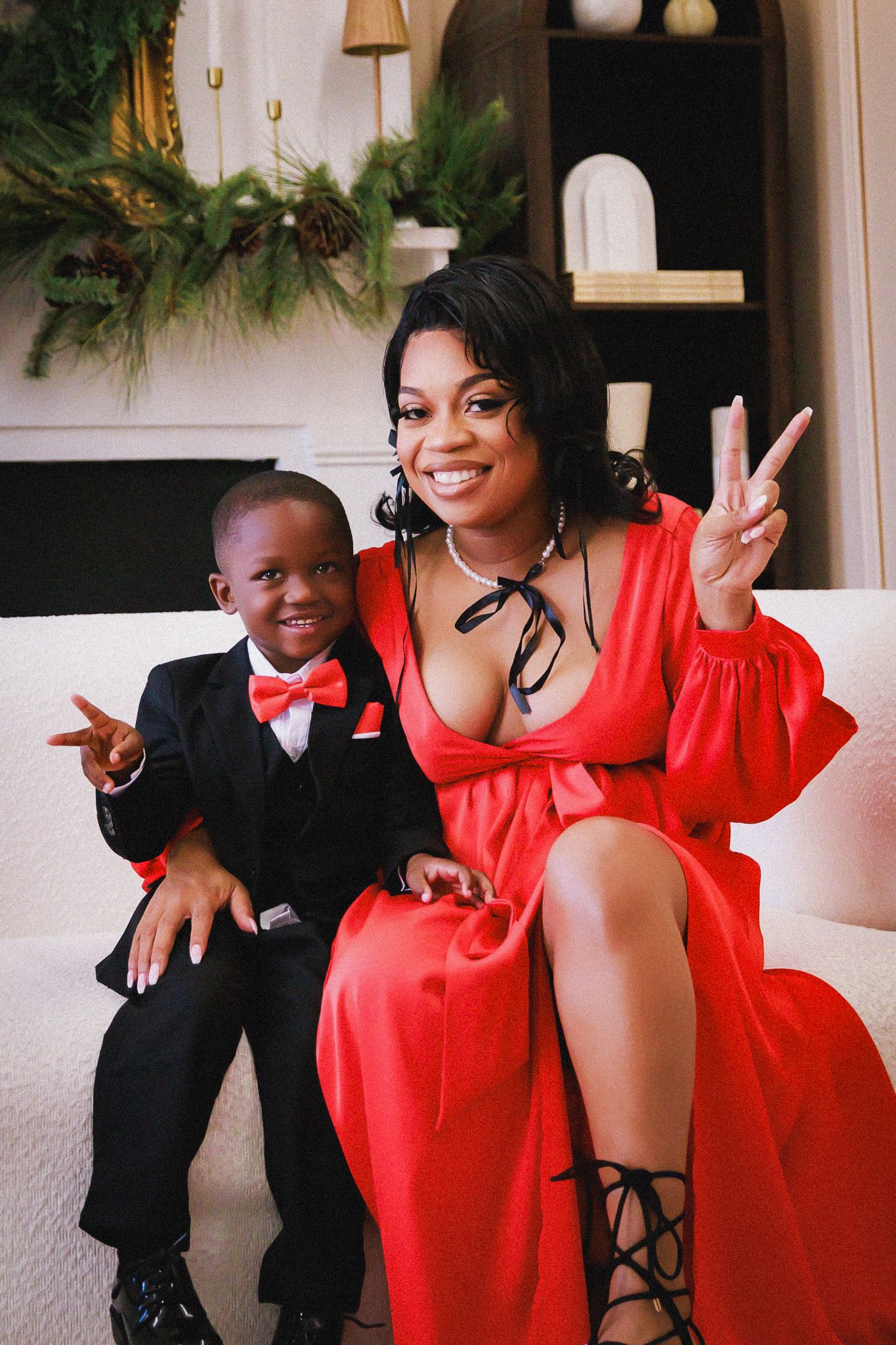 A parent and child in festive attire pose together on a couch, making peace signs in front of holiday decorations.