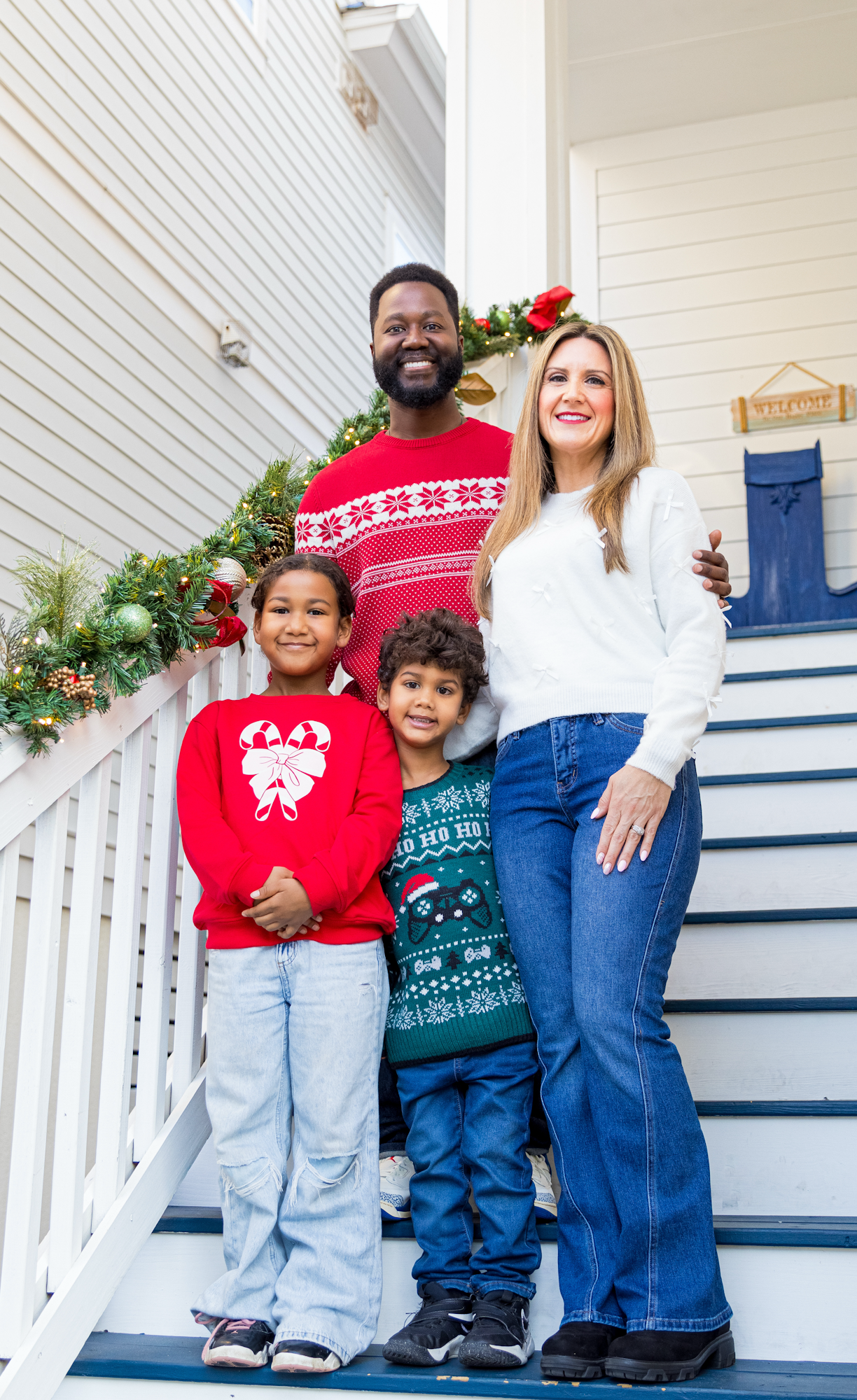 Family wearing festive holiday sweaters poses together on white porch steps for Christmas photo.