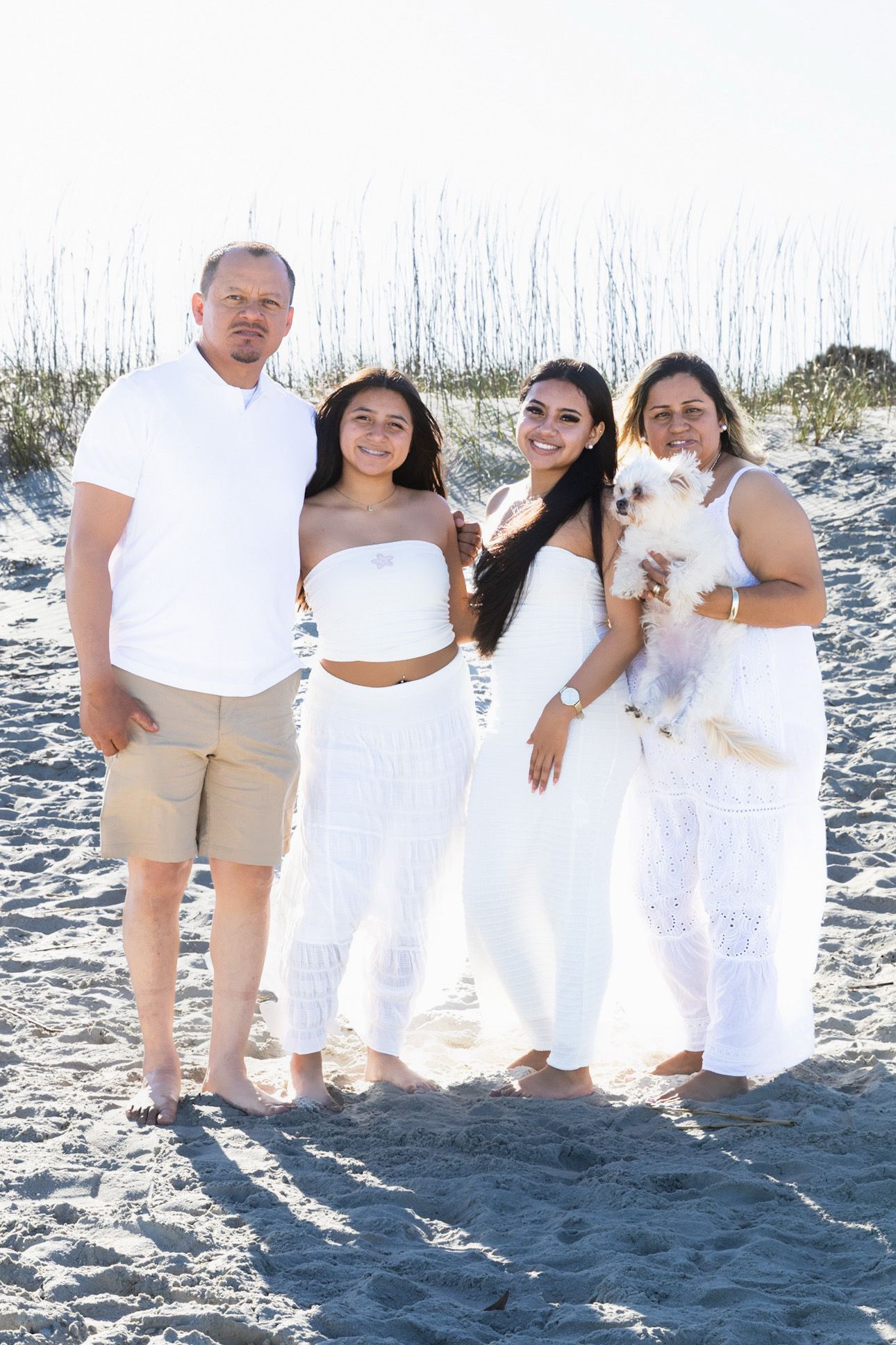 Family of four in white outfits posing with a small dog on a sunny beach with sea oats in background.