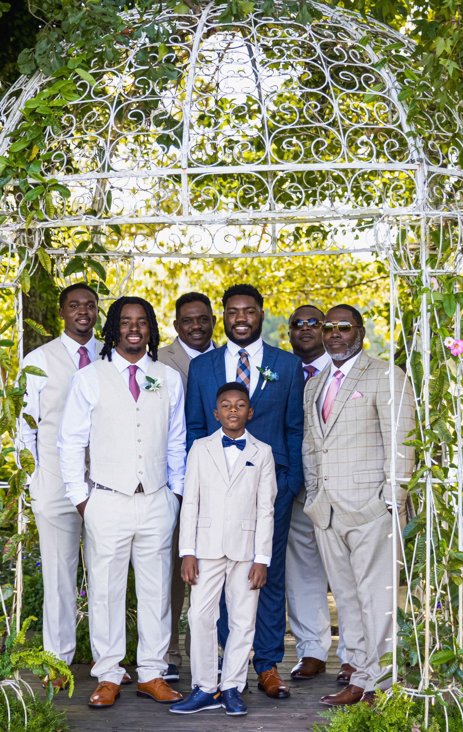Groomsmen in light suits pose under decorative white gazebo with greenery.