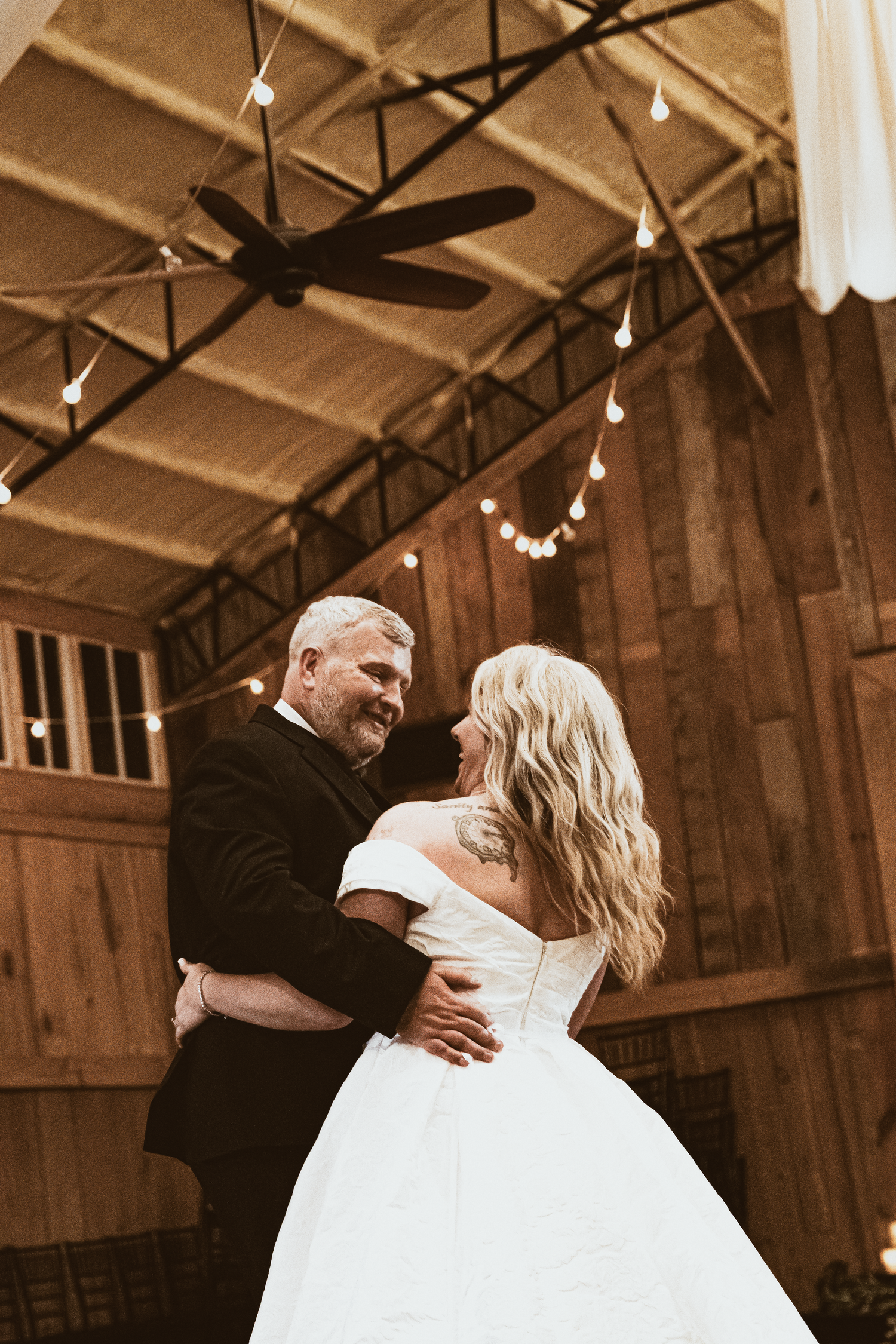 Bride and groom share first dance in rustic barn venue with string lights and wooden ceiling.
