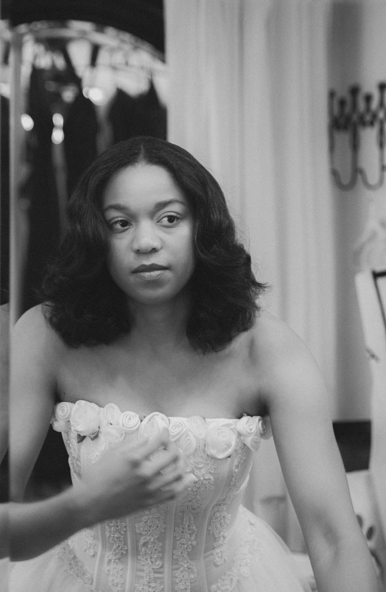 Black and white photo of a young woman in a strapless floral lace bridal gown posing in a bridal boutique.