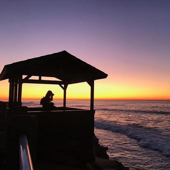 Panoramic ocean sunset with silhouetted beach structures against purple sky.