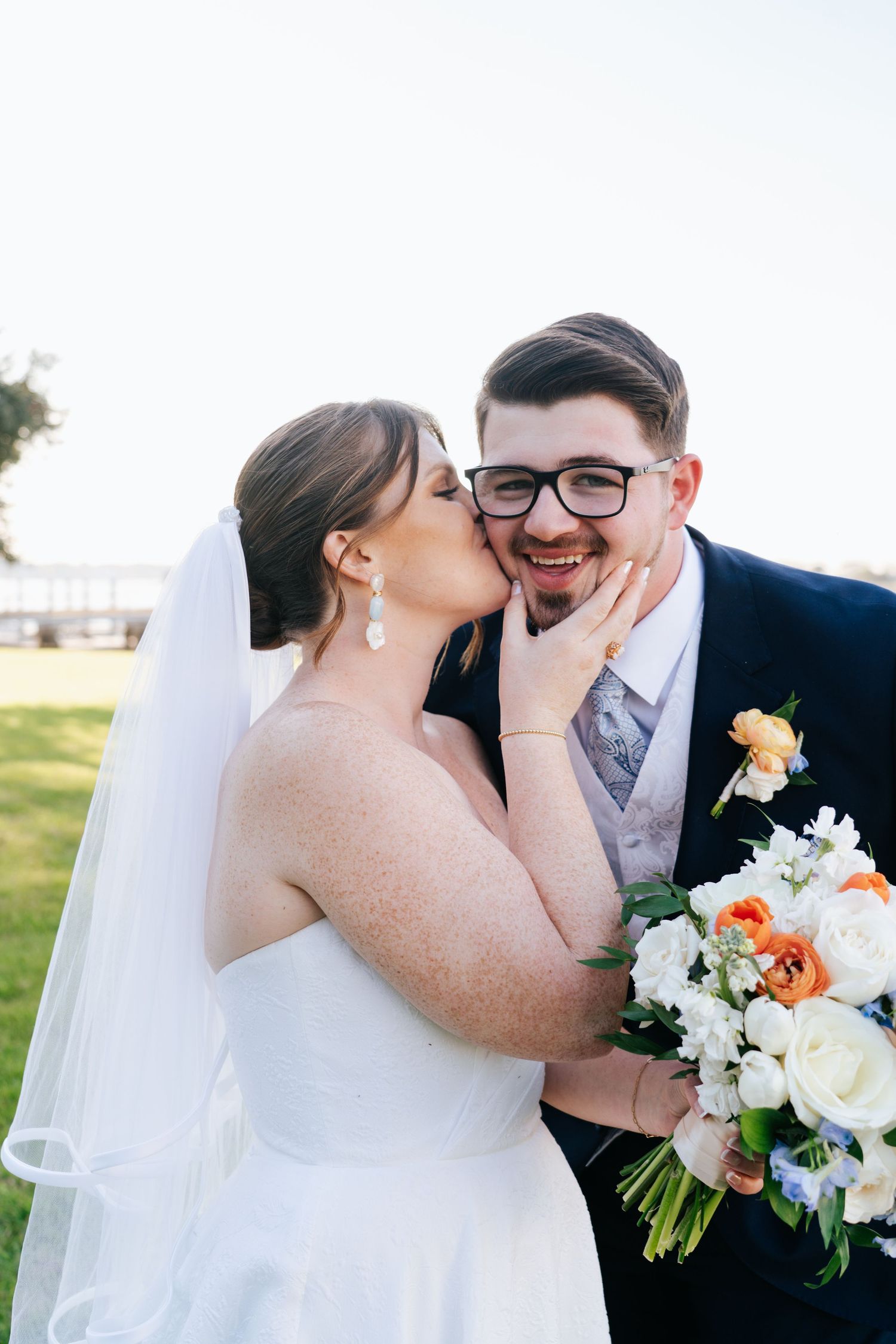 Newlyweds share romantic kisses while holding a bouquet of white and orange flowers.
