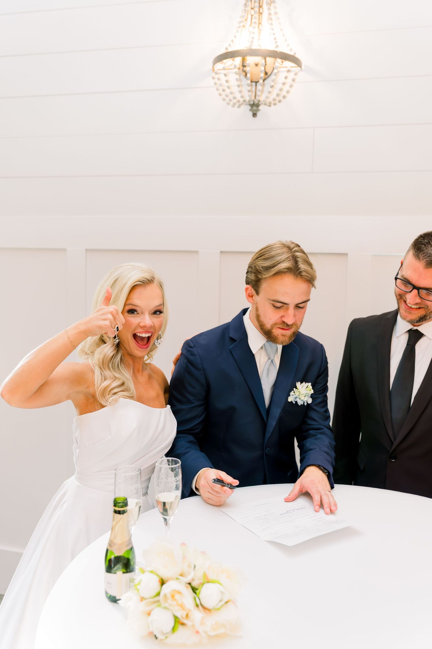 Excited bride makes peace sign as groom signs wedding certificate with officiant at white decorated table.