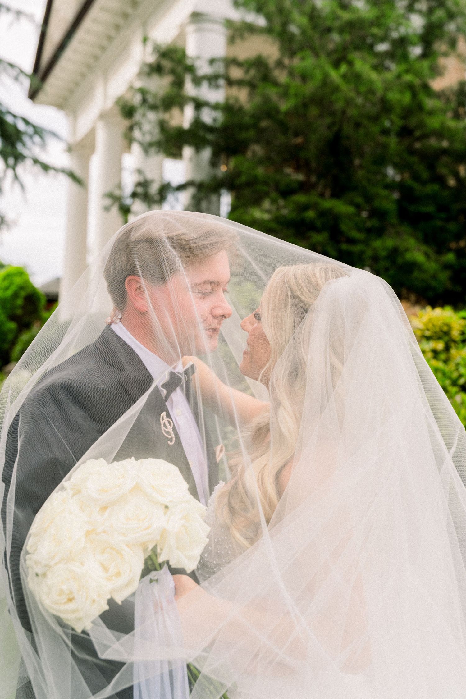 Bride and groom share romantic moment under flowing white veil, bride holding white rose bouquet outdoors.