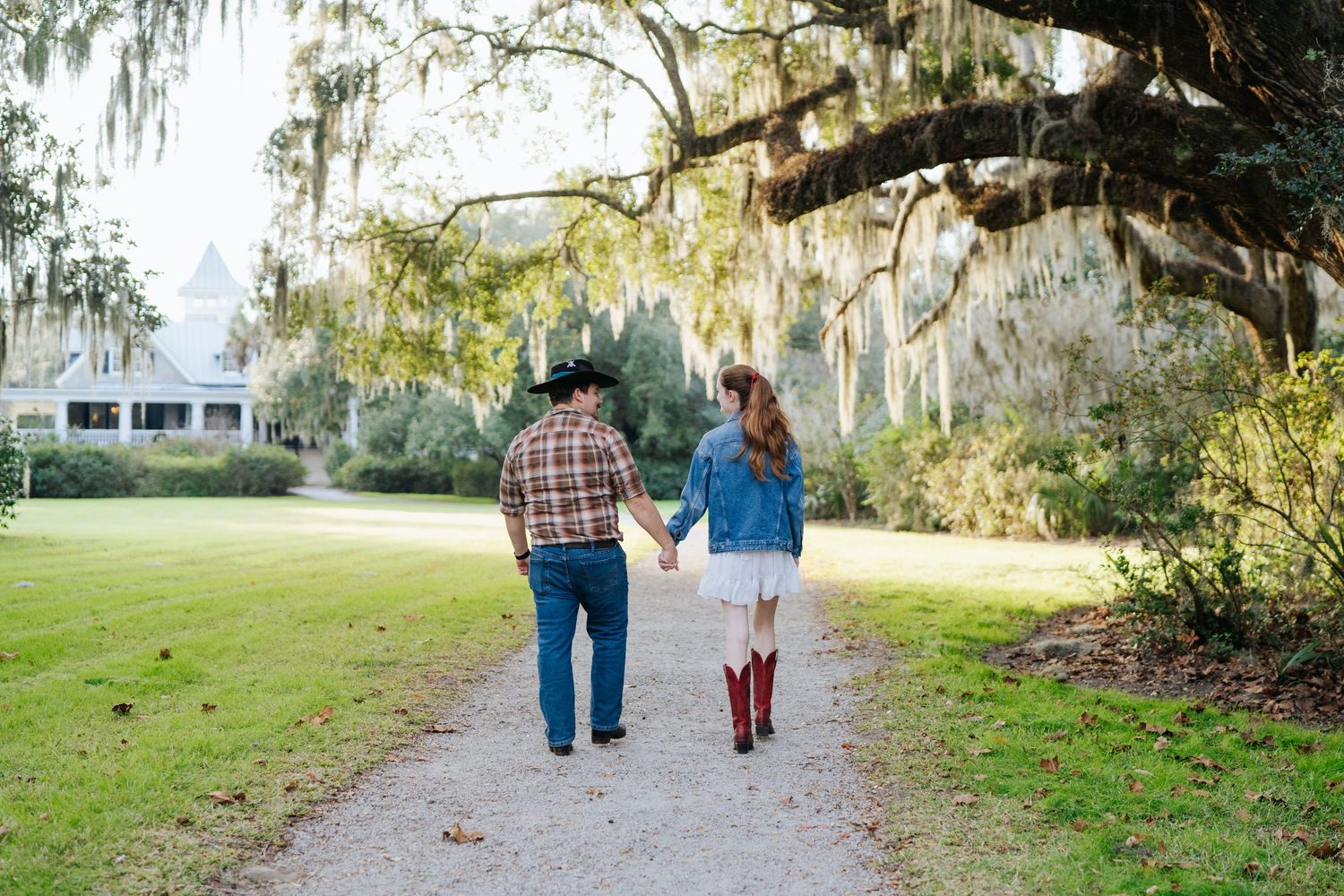 Couple walking hand in hand down a path under Spanish moss covered trees.